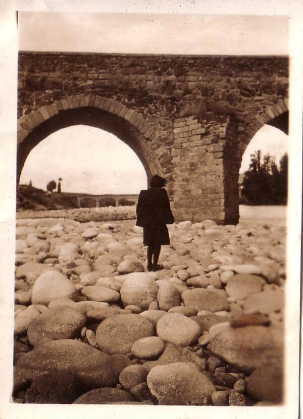 En la imagen, una mujer vestida de negro resiste al viento sobre el lecho seco de un río con los arcos de lo que parece un puente romano (o al menos románico) al fondo.